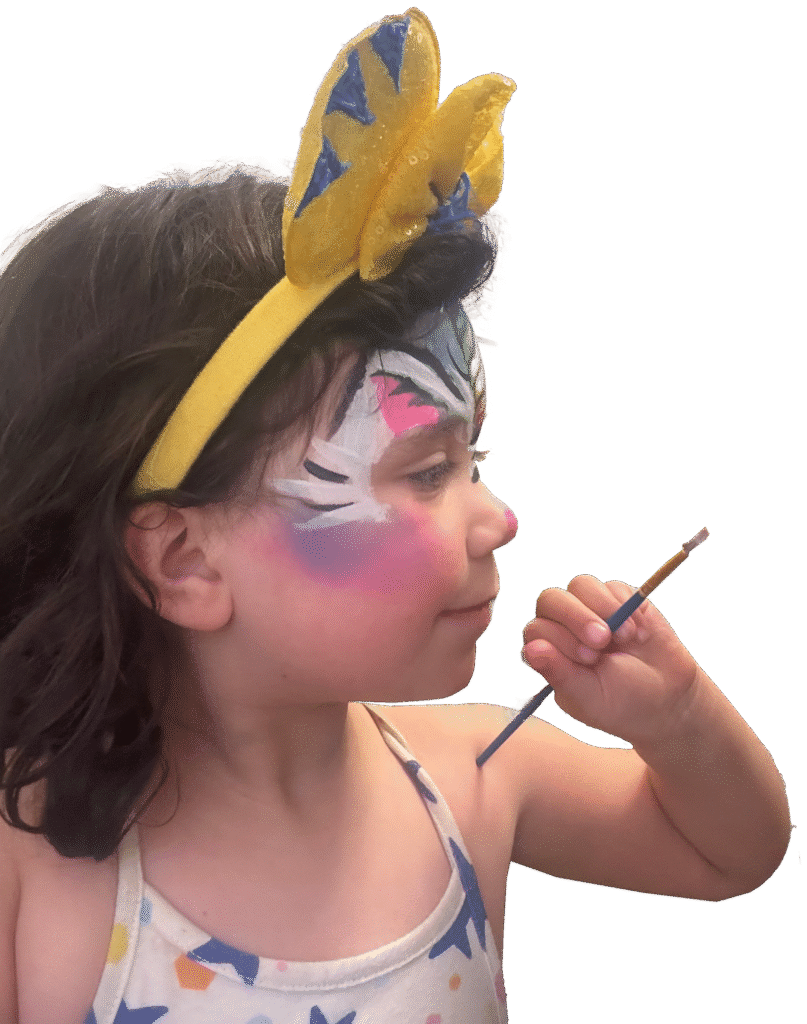 Girl with her face painted like a unicorn holds up a paintbrush as though painting.